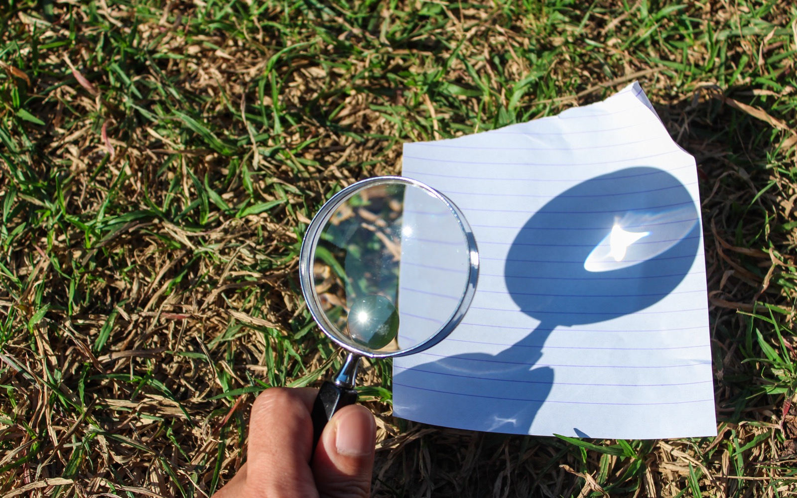 A person holds a magnifying glass and focuses sunlight through the glass to create a small dot of light on a piece of paper.
