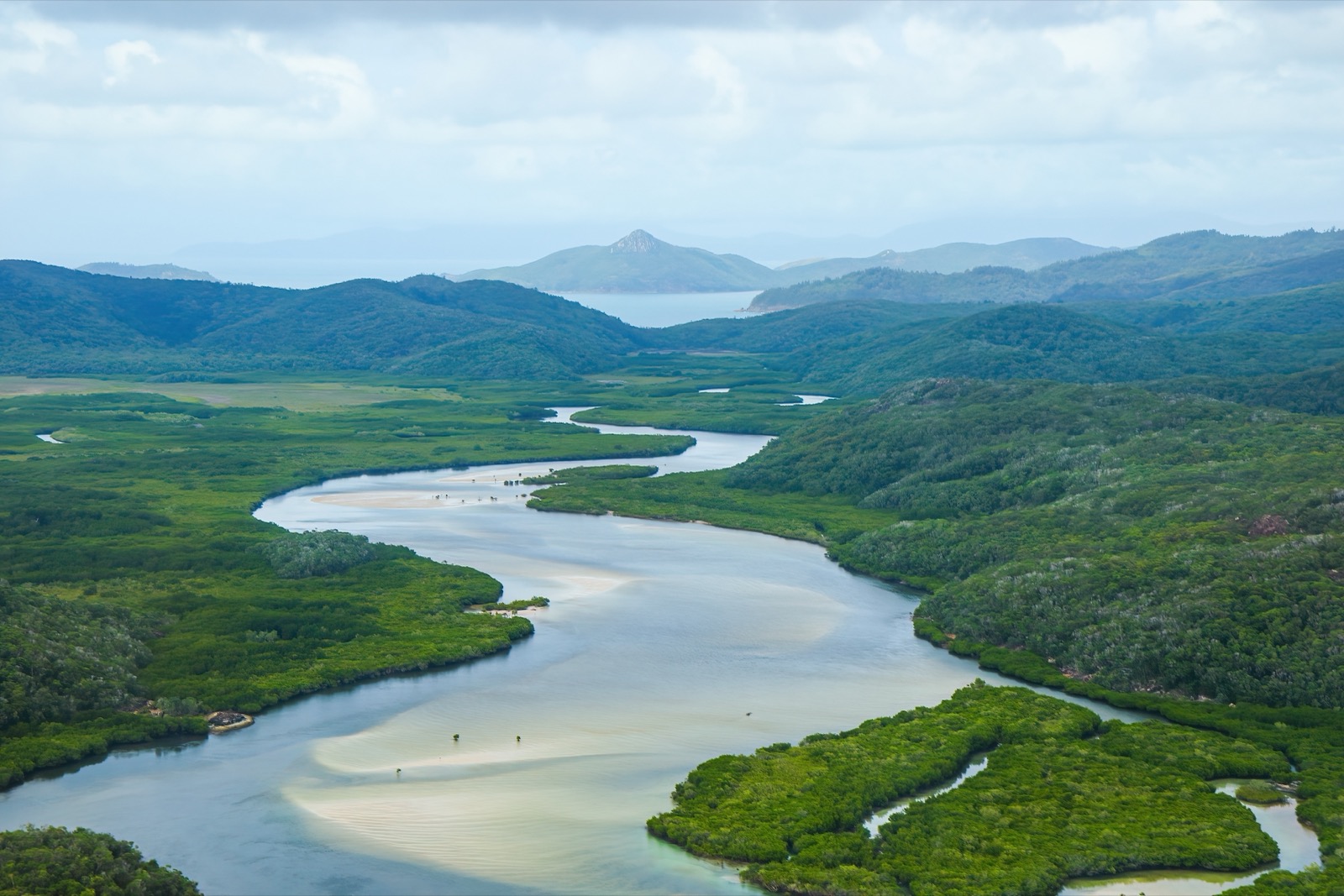 An aerial view of a wide, winding river flowing through dense mangrove forests toward a distant coastline, with green hills and mountains in the background under a partly cloudy sky.