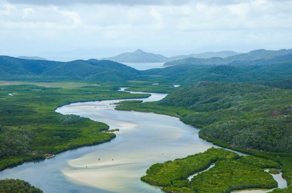 An aerial view of a wide, winding river flowing through dense mangrove forests toward a distant coastline, with green hills and mountains in the background under a partly cloudy sky.