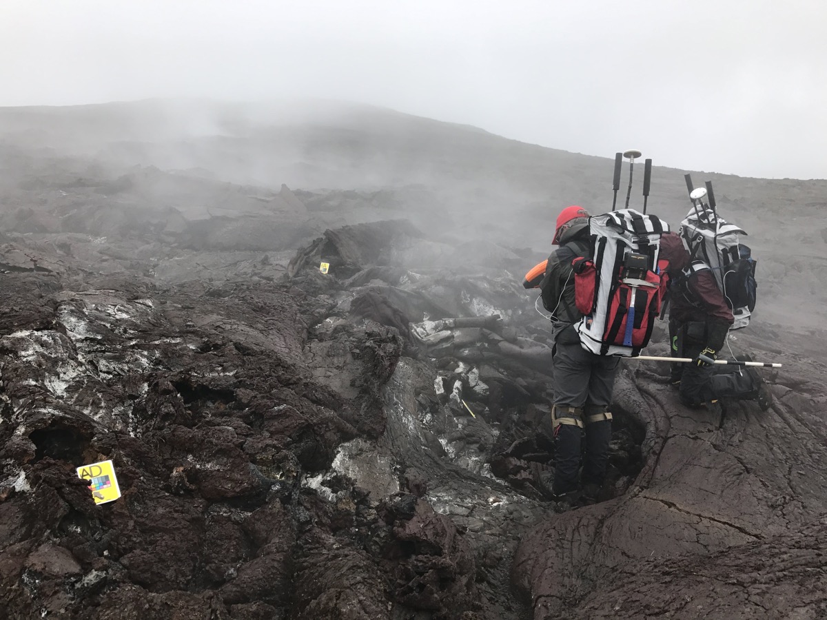 two people fully clothed in extreme hiking gear with large backpacks with antennas coming out stand on a black, molten landscape with pockets of steam seeping from the ground