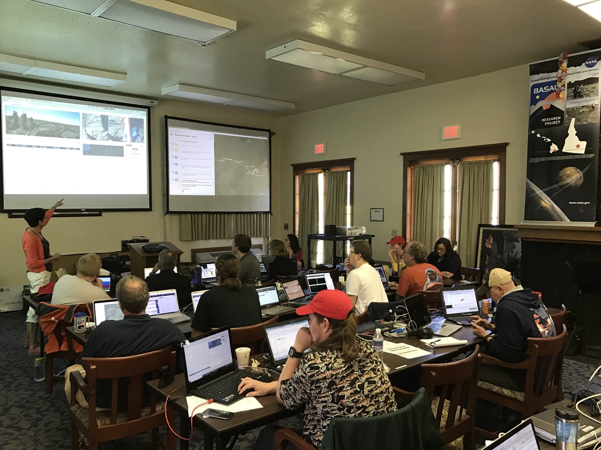 A dozen or so people sit in a room at three rows of long desks with computers. at the front of the room is an instructor gesturing at two projected images on the wall