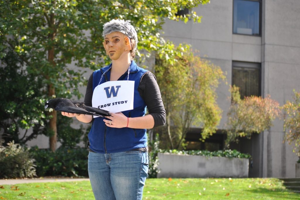 researcher with mask and gray wig holds dead crow