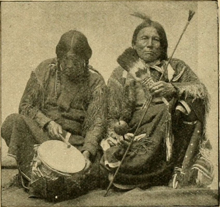 a black and white photo of two kiowa native americans with instruments sitting on the ground