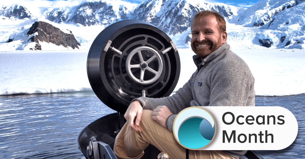 a man sitting on top of the submarine in the water with snowy mountains behind him