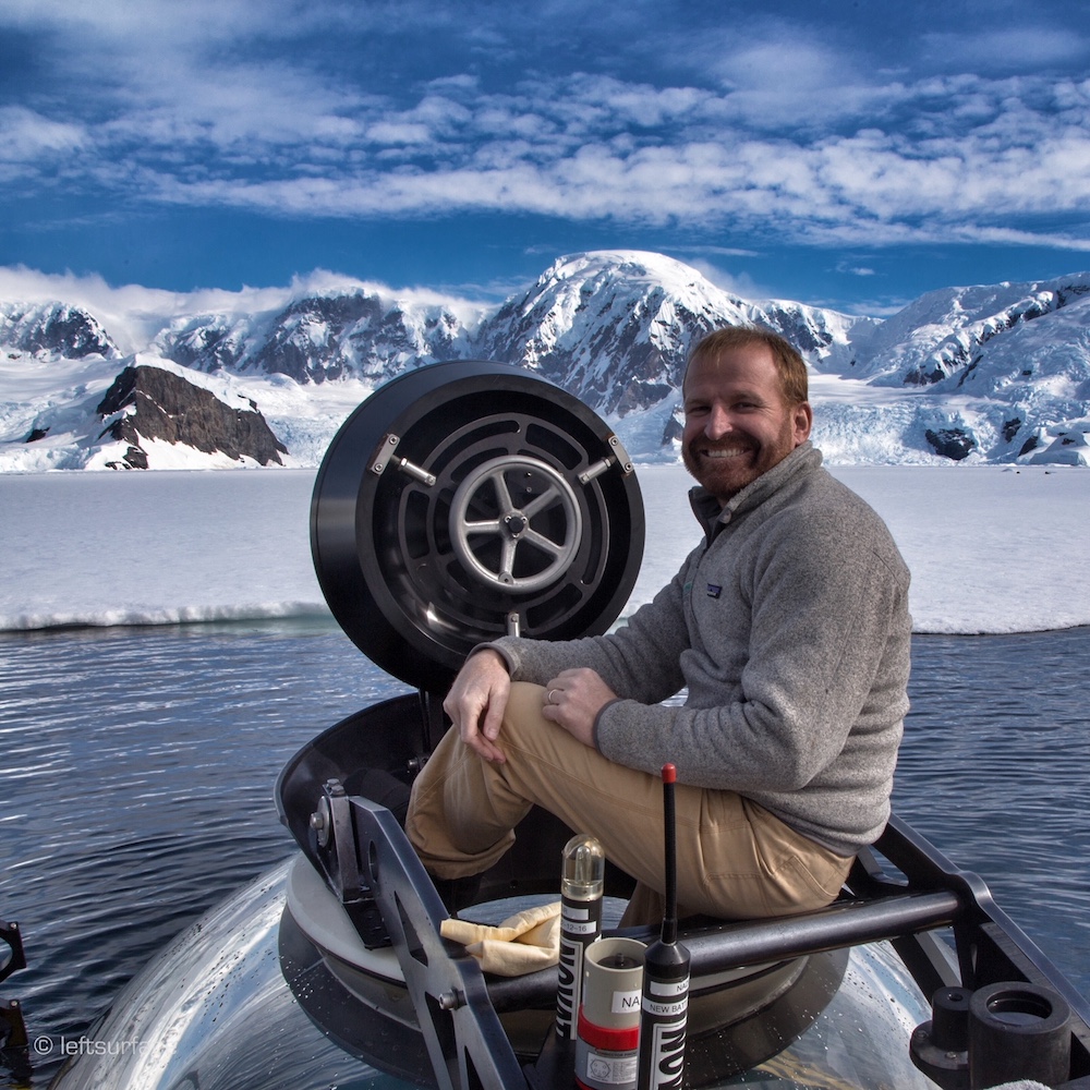 a man sitting on top of the submarine in the water with snowy mountains behind him