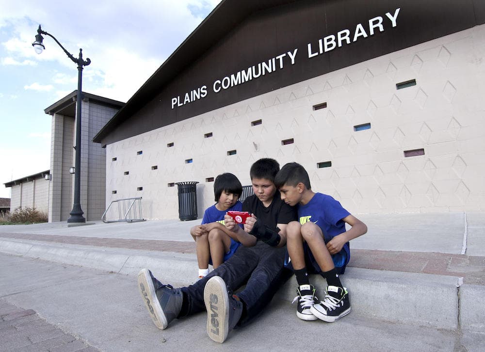 three children sitting in front of a library playing a handheld video game