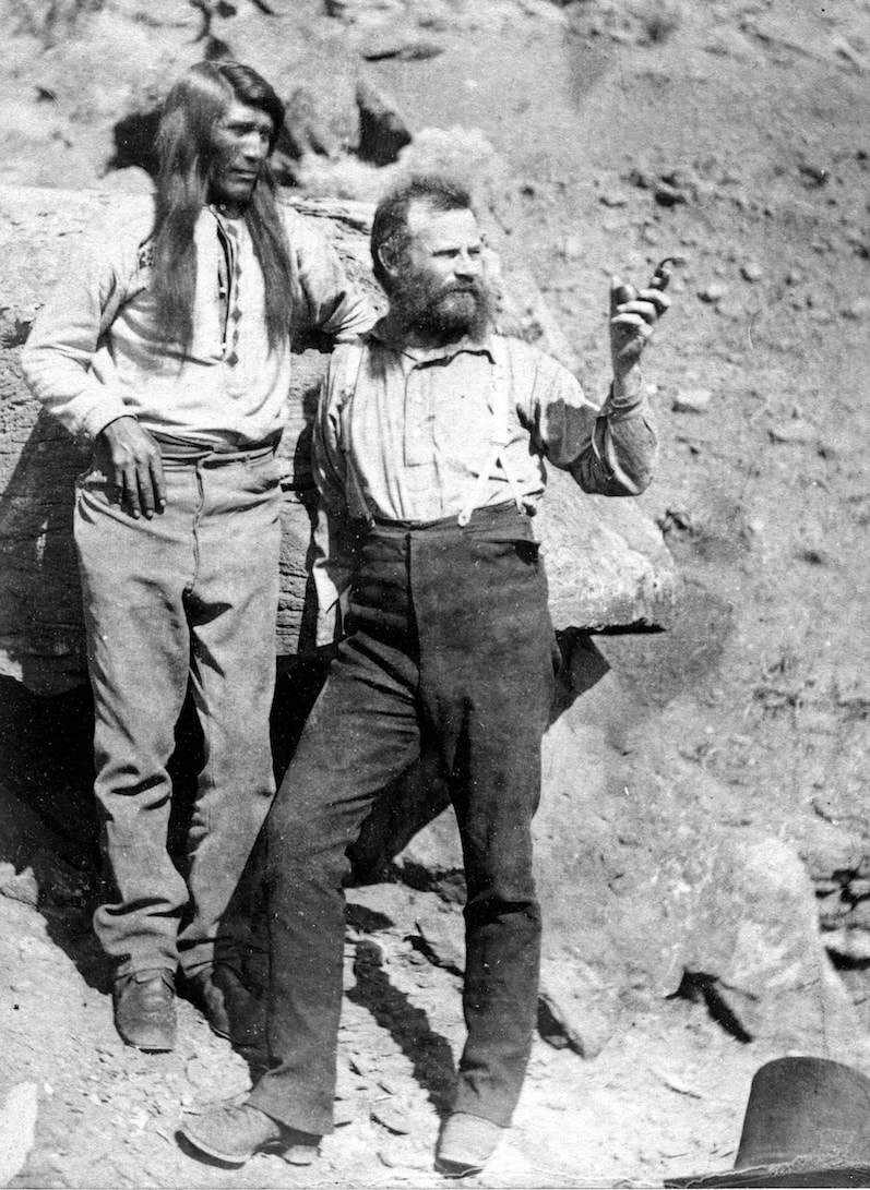 a black and white photograph of two men standing next to each other against a rock, one bearded (powell) pointing his finger ahead of him