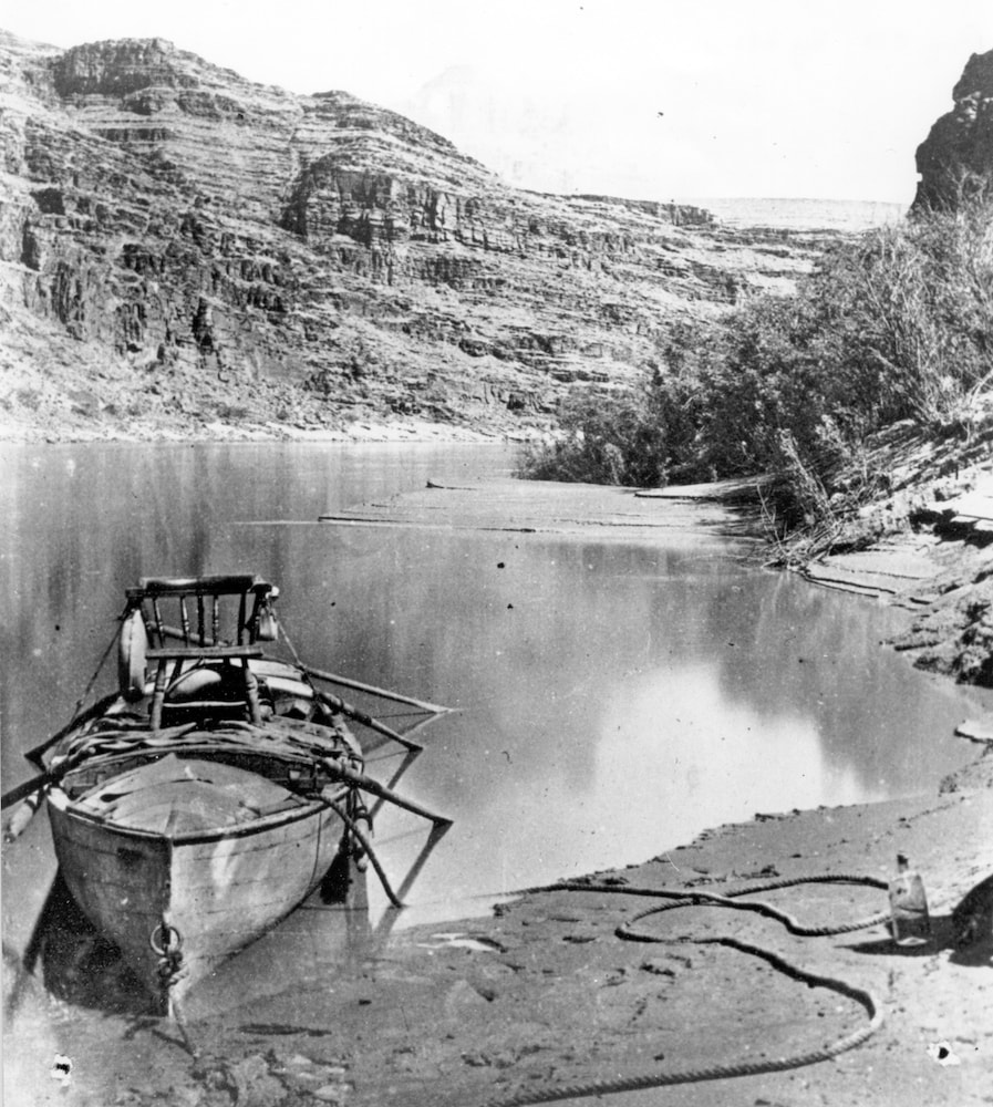 a closer view of powell's boat with the chair strapped to it, docked on the shore of the river