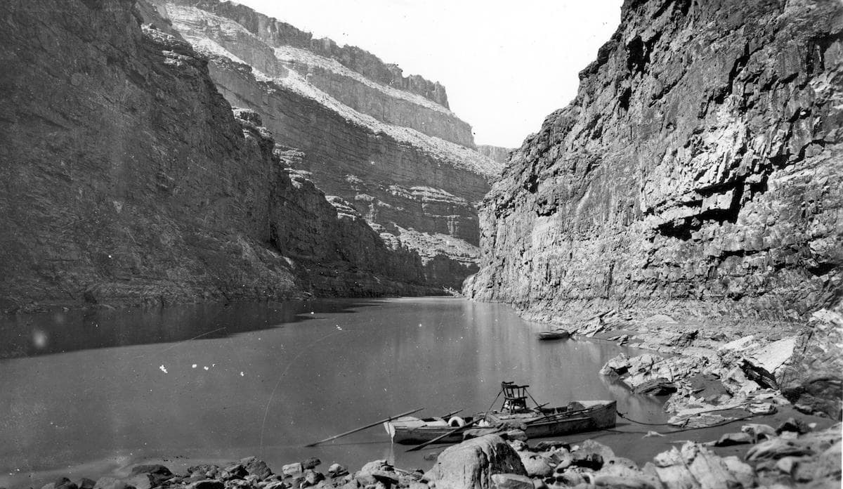 a black and white photograph of a boat docked on the shore of the colorado river amidst the sweeping landscape of the canyon