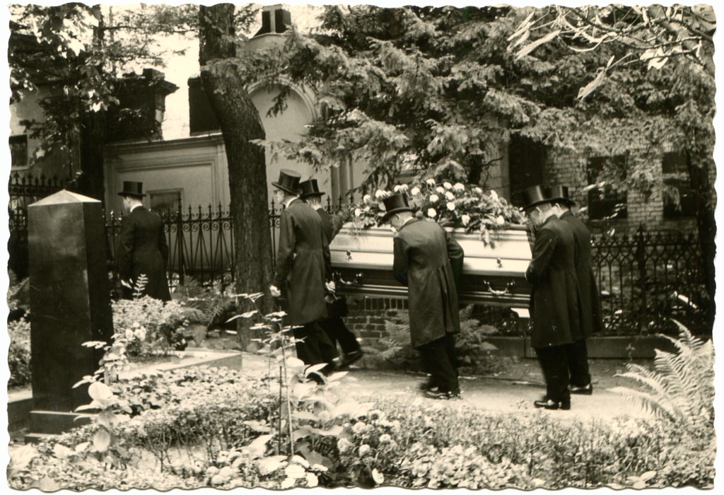 An antique photo shows a group of men in black overcoats and bowler hats, carry the coffin with the deceased at the cemetery