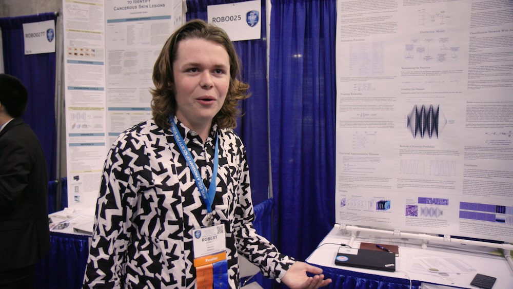 a young male student with shoulder length brown hair and a patterned black and white shirt in front of a science fair board