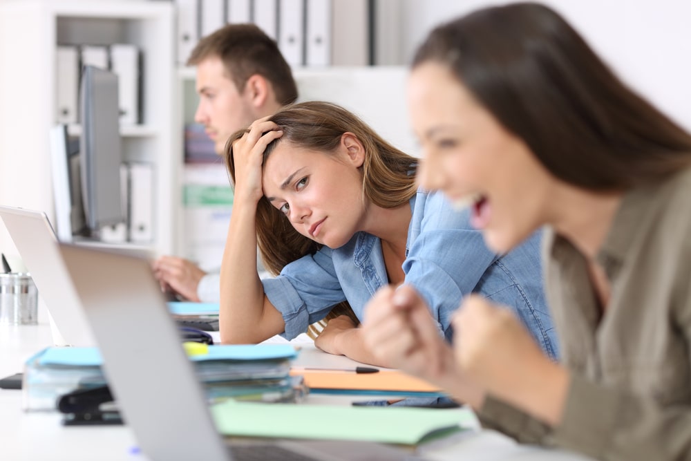 Worried worker beside a successful one who is excited reading good news on line at office