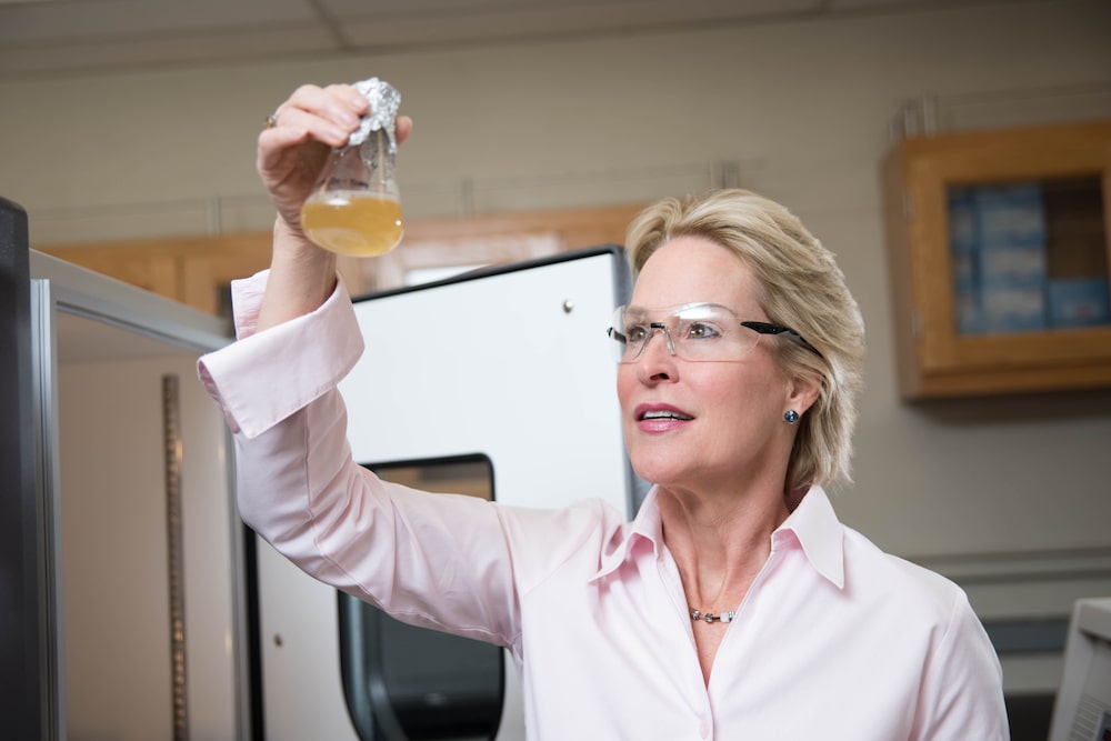 a woman in a lab wearing safety glasses holding up a beaker filled with liquid up to the light