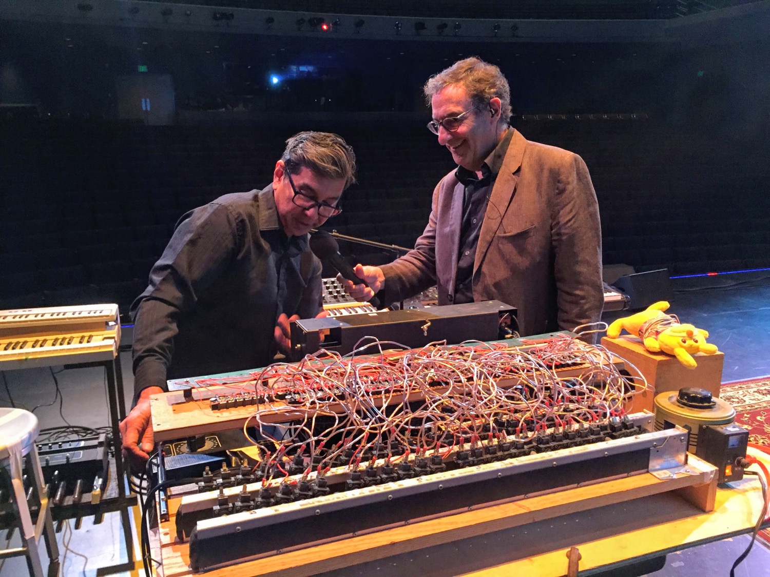 two men, one holding a microphone up to the other who is gesturing to a machine with many wires coming out of it