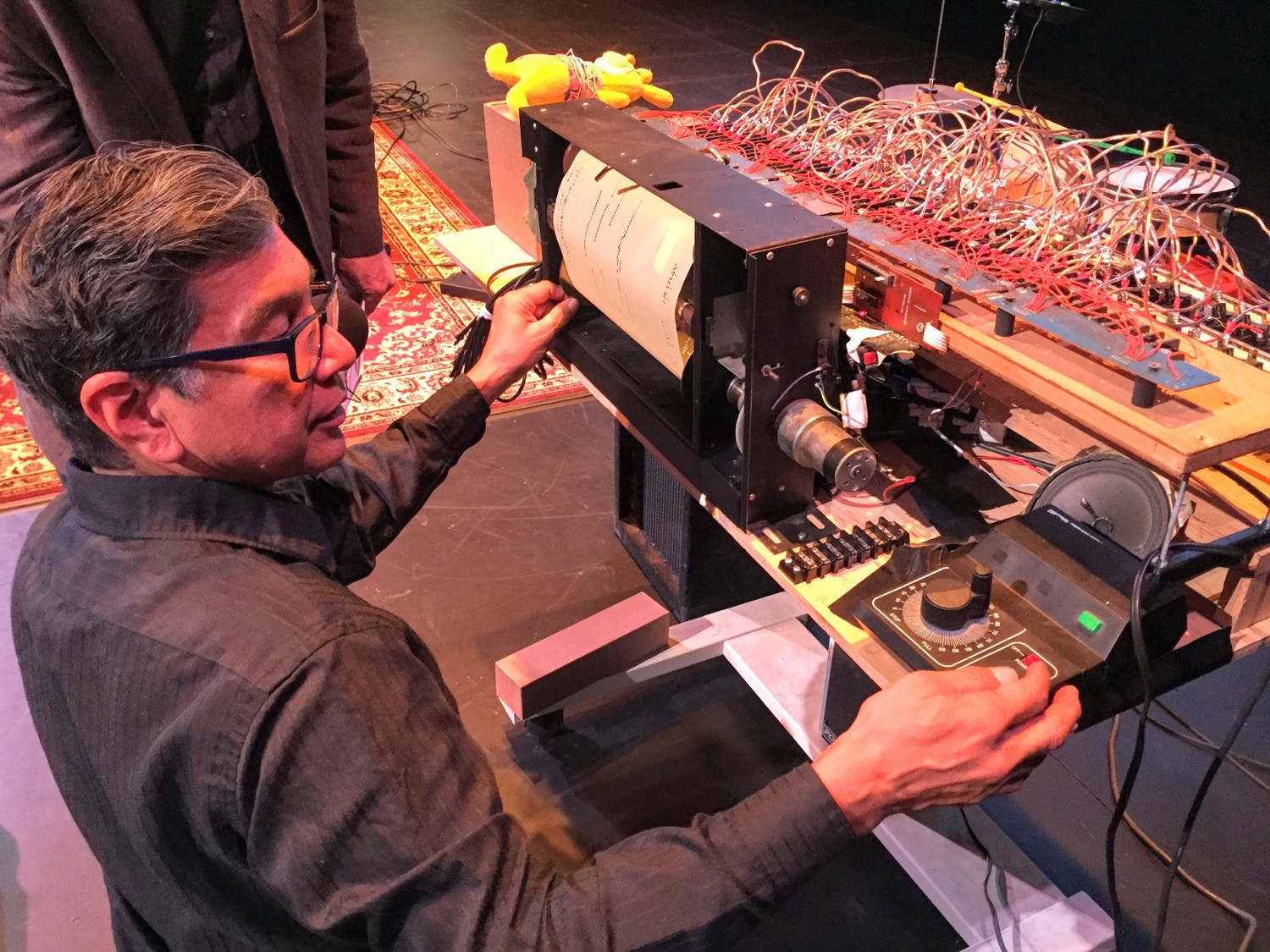 a man crouches at the back of a complicated looking machine with dials and many wires