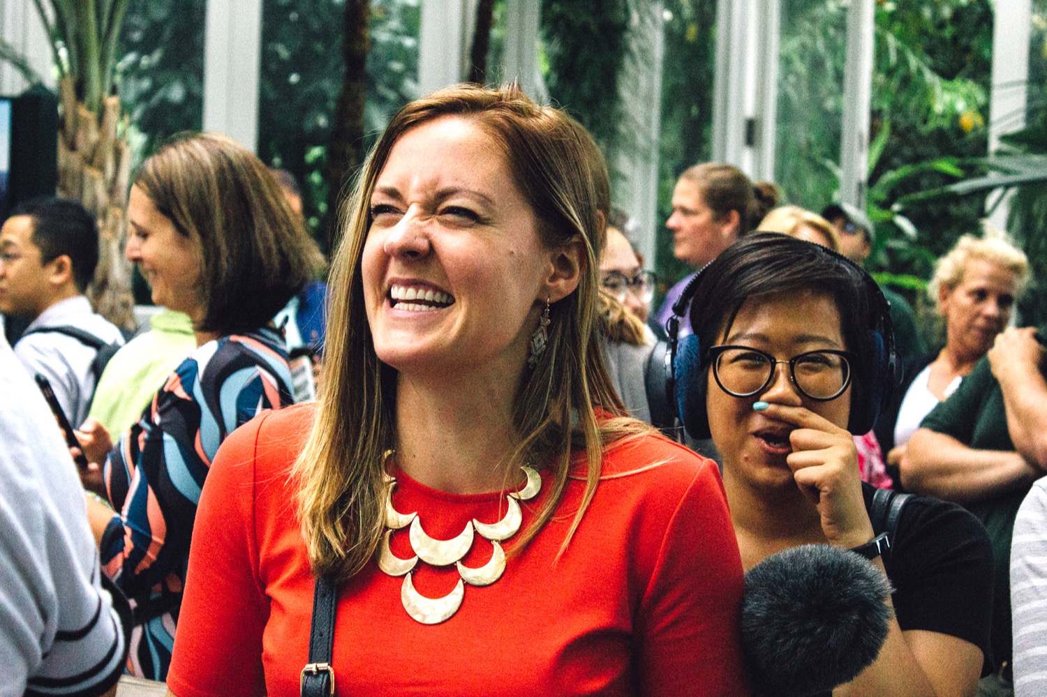 in a crowd of people, there are two women, one who's nose is wrinking and smiling, and the other, wearing headphones and carrying a microphone, is putting a finger under her nose