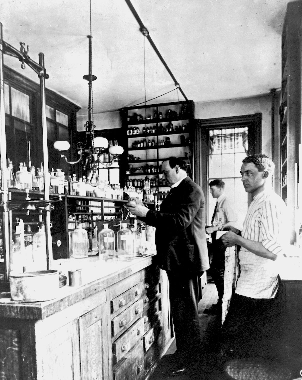 a black and white photo of a chemistry lab with a man in a coat holding equipment and two other men who are assistants