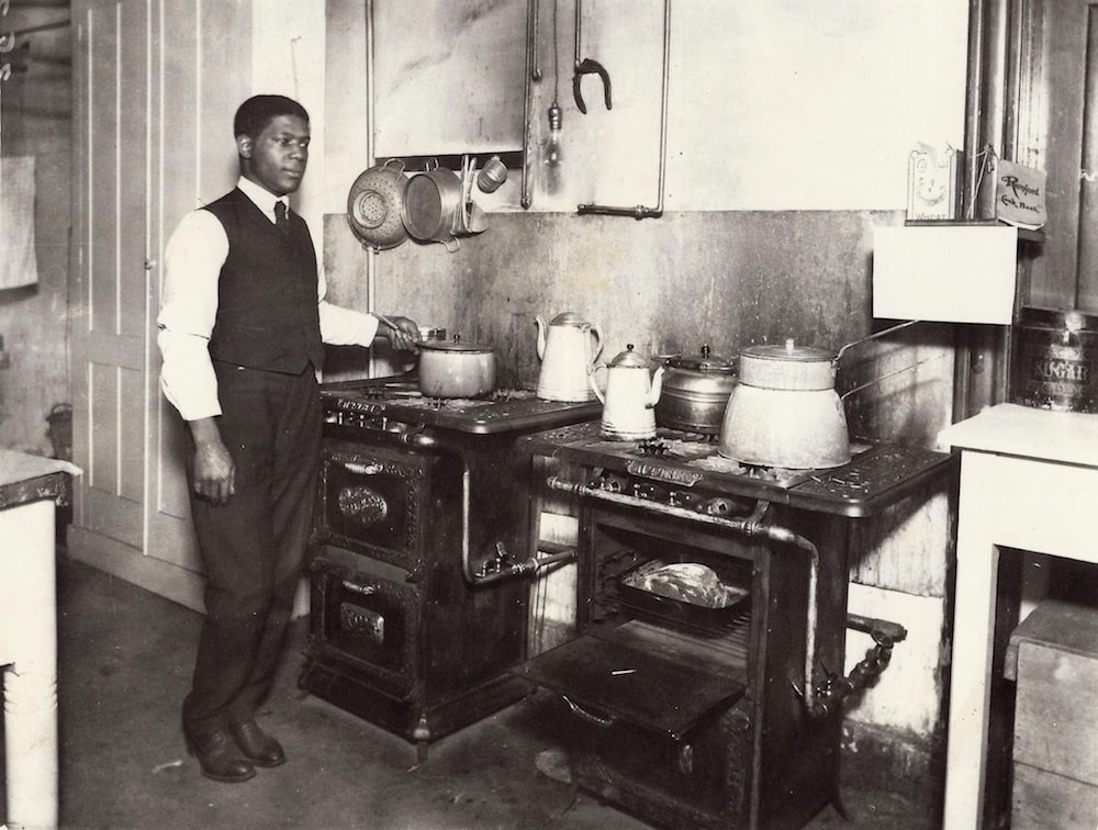 an african american man in a vest and tie holding a pot in a kitchen