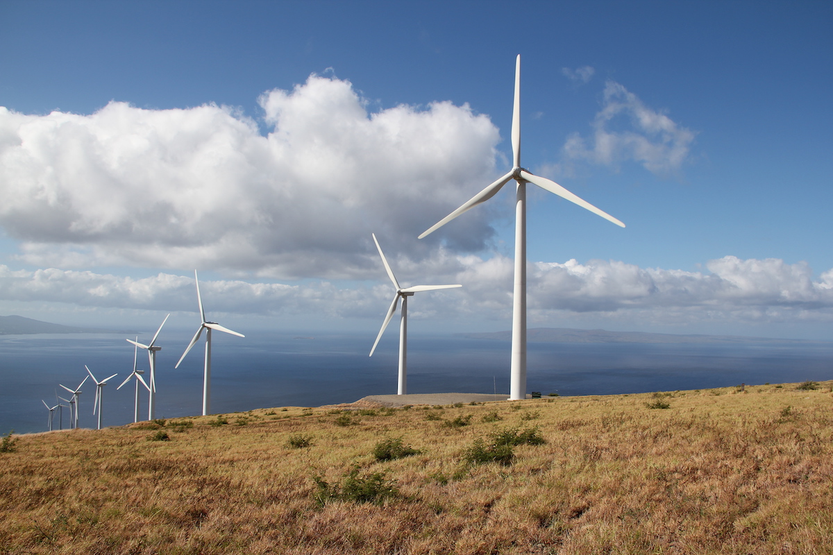 four wind turbines in a row on a ridge overlooking the ocean and a blue sky