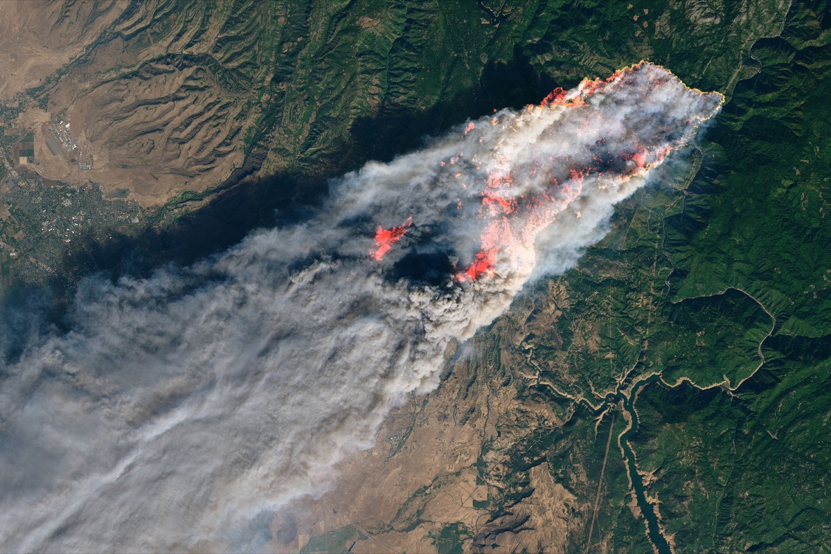 an satellite photo of a massive fire with a large trail of smoke and bright orange outlines indicating the edges of the fire