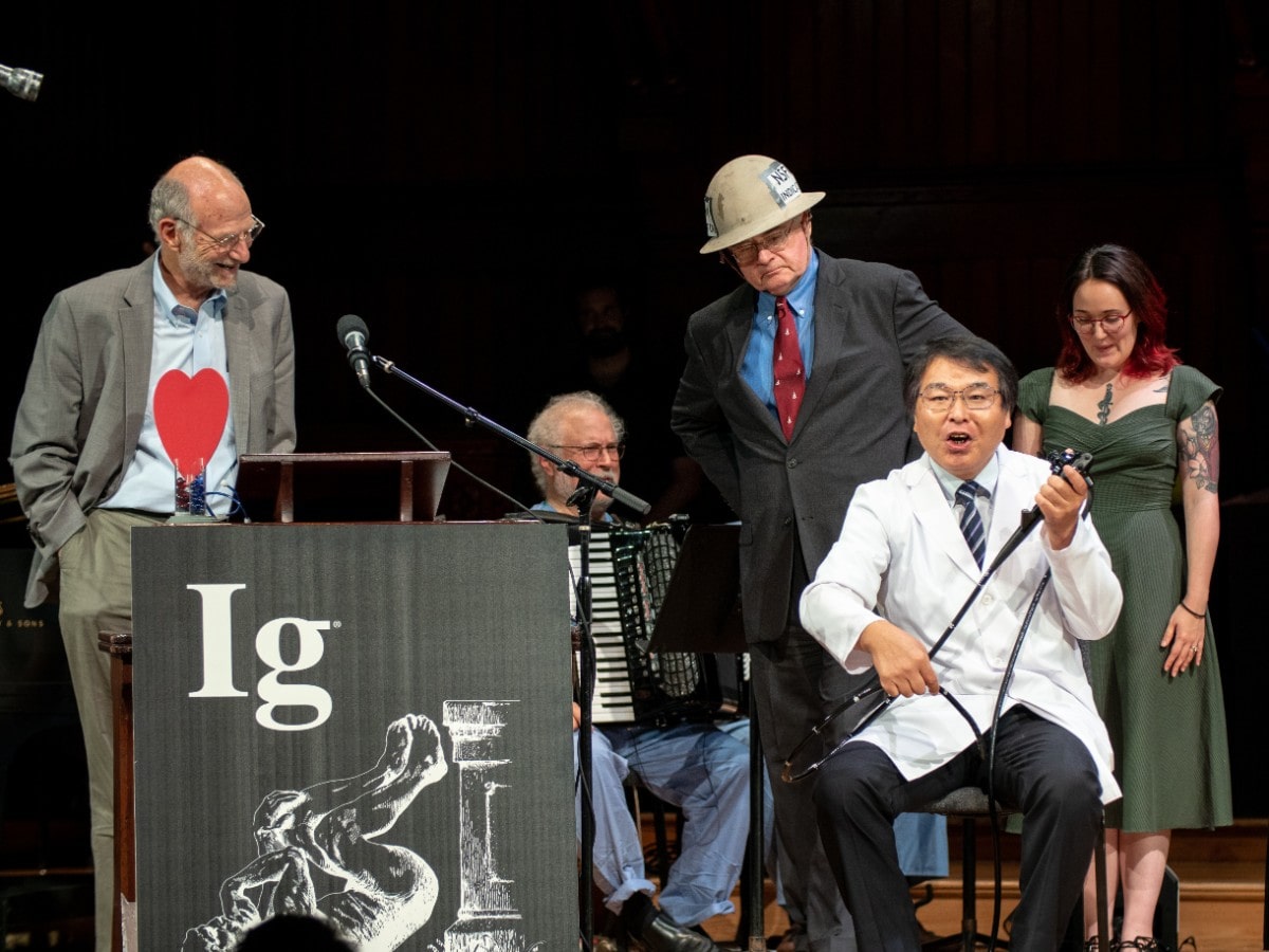 a man in a lab coat holds medical equipment and sits in a chair with an anguished face. two other men are on stage listening to him