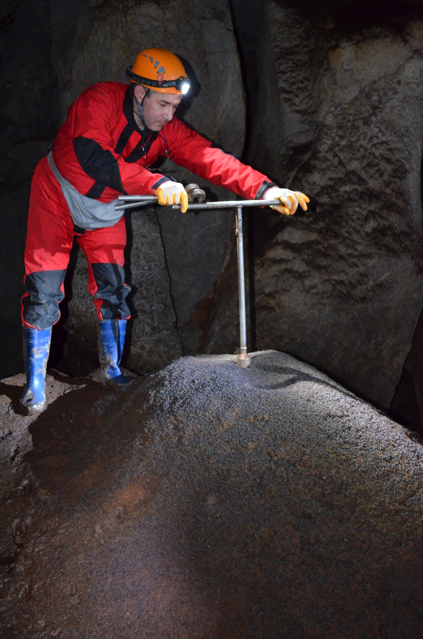 a man in an orange safety jumpsuit with a helmet that has a light putting a T-shaped metal device into knee-high hill in front of him