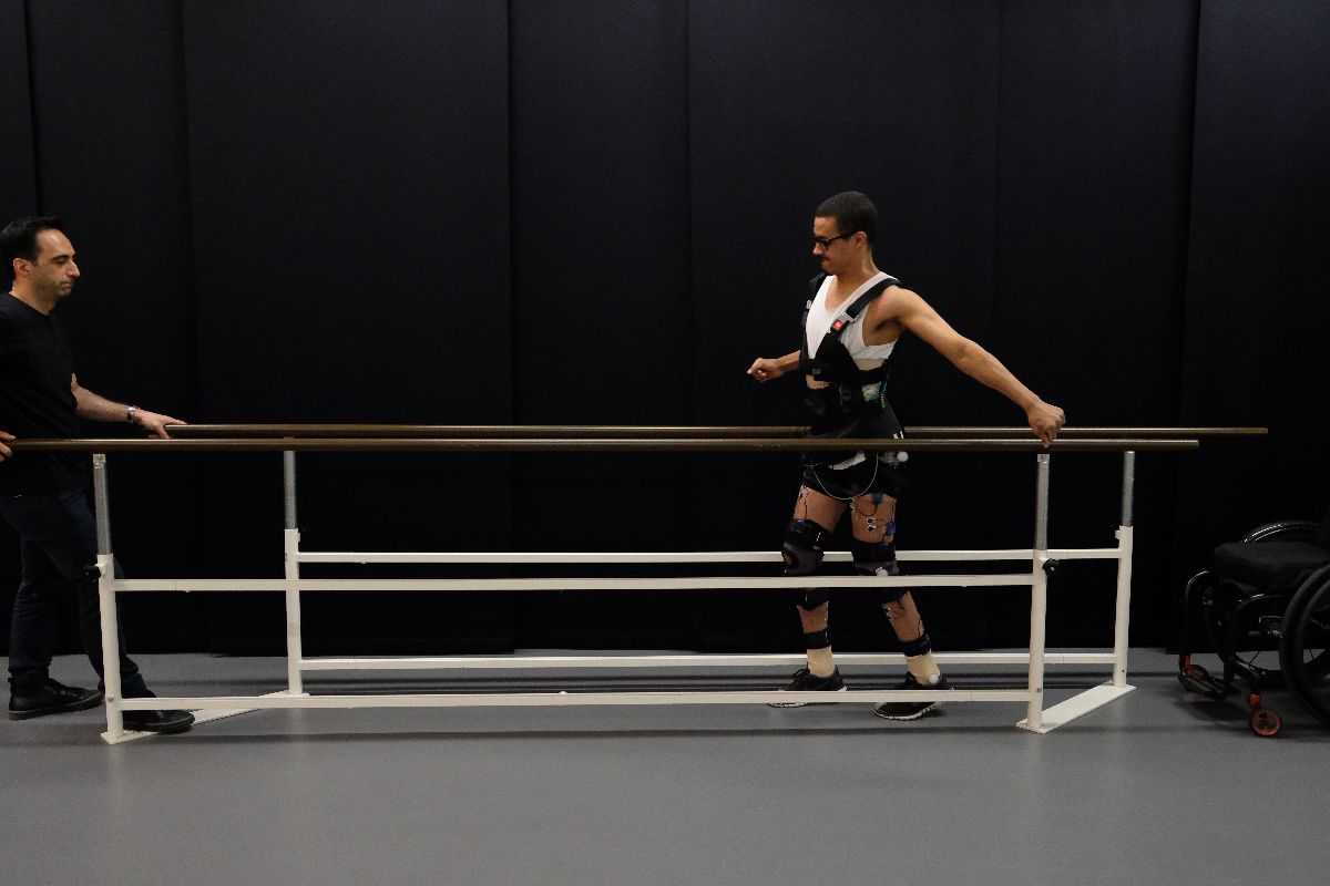 a man wearing research equipment around his waist and on his legs walks with his hand on a ballet bar