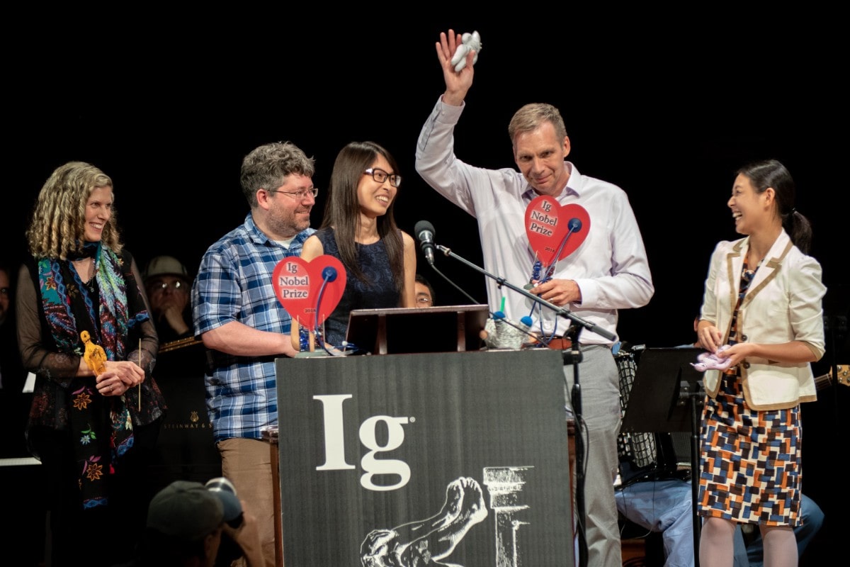 an older man receives an award and makes a speech at a podium while holding up a little vodoo doll.