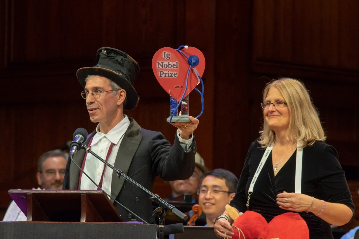 a man in a top hat holds an award made of a cutout heart with a stethoscope around it, that reads "ig nobel"