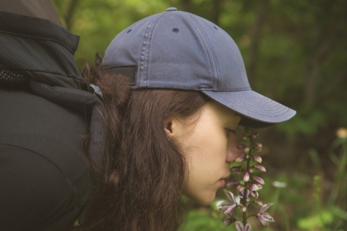 a woman with long brown hair and wearing a baseball cap smelling flowers in a forest