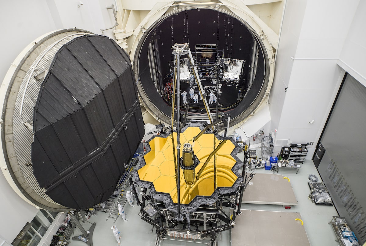 an overhead shot taken in a cavernous room. one wall is a giant open hatch that has to be at least 30 feet in diameter. the telescope, with its large, golden hexagon mirror, emerges with technicians, tiny in comparison, working around it
