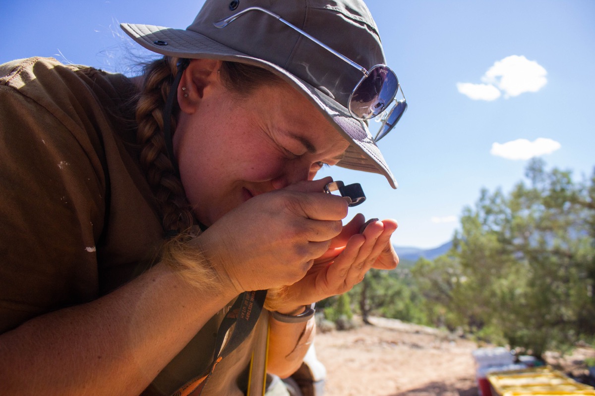 a female paleontologist uses a magnifying glass to look at a sample at a campsite in the desert