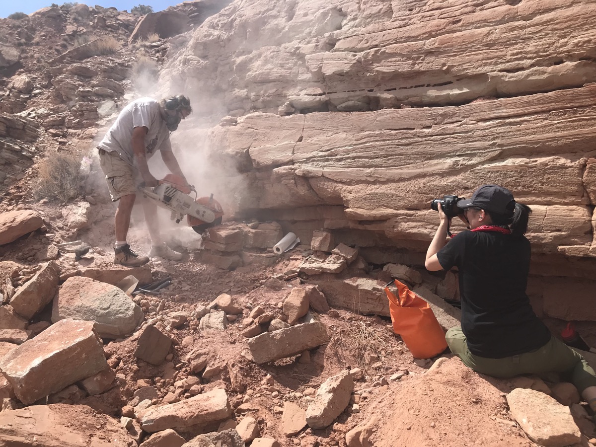 a woman crouches down next to a cliff in the desert taking a photo of a man rock sawing 