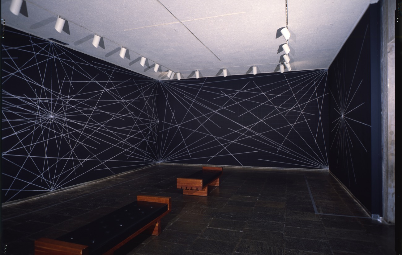 photo of a room with three walls and two benches in the center, on the walls are a collection of white lines, emerging from a single point. there are several of these arrangements on the walls