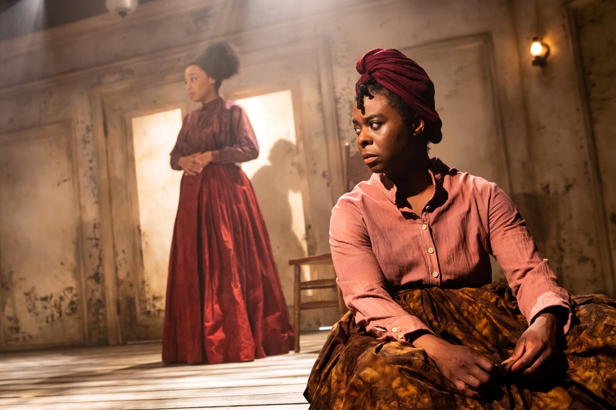 an intense moment with two women of color on the stage. one sits in the foreground looking off into the distance, while the other stands in the background with her hands over her stomach