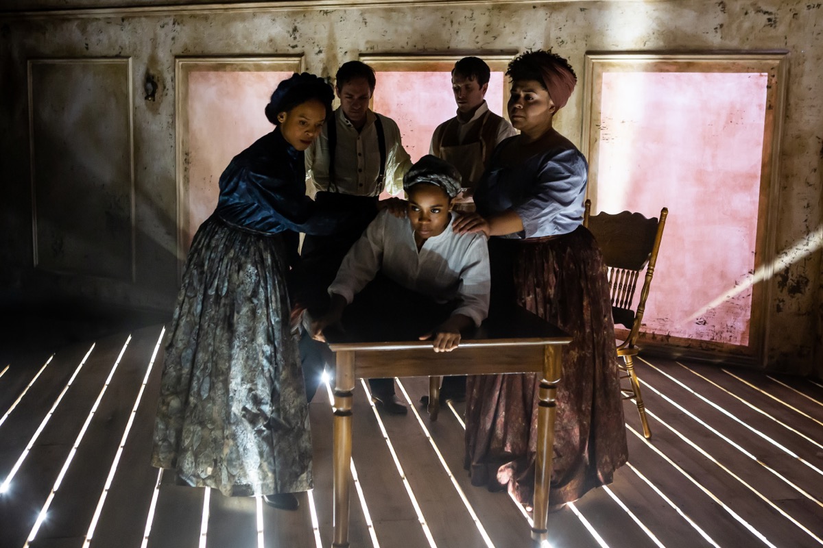 five people around a table on a stage with dramatic lighting shining through slats of the wooden floorboard. two women stand on the left of the table assisting a black woman lying on her belly on the table, while the two men are medically examining her