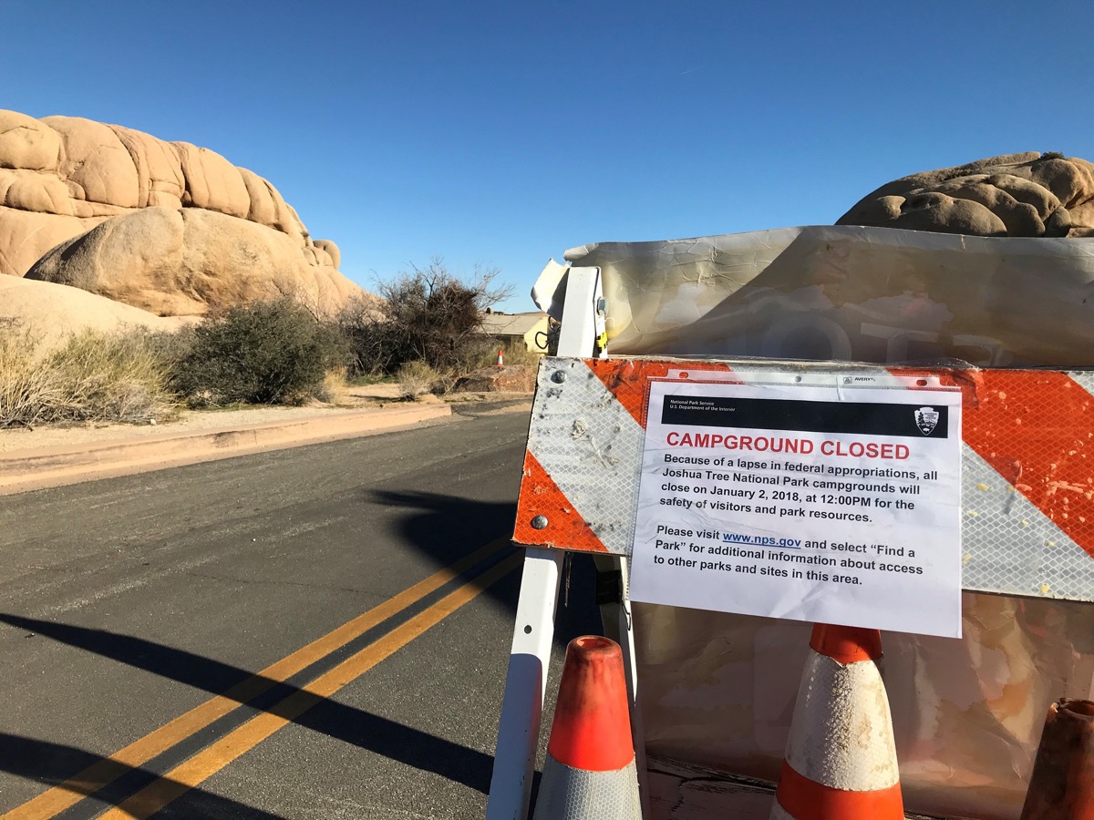 a road sign that says "campground closed because of a lapse in federal appropriations, all Joshua tree national park campgrounds will close on January 2, 2018, at 12:00 pm for the safety of visitors and park resources. Please visit www.nps.gov and select find a park for additional information about access to other parks and sites in this area." the sign is on a road next to large sandstones in the park