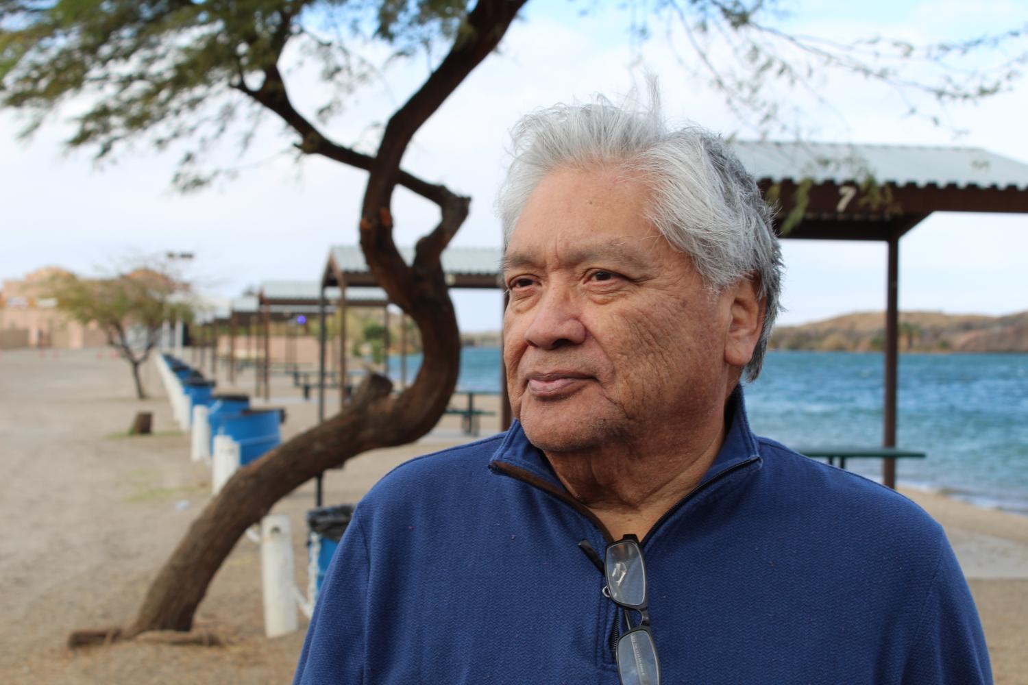 an older man looks off into distance, in the background is a lake and a dozen or so small shades