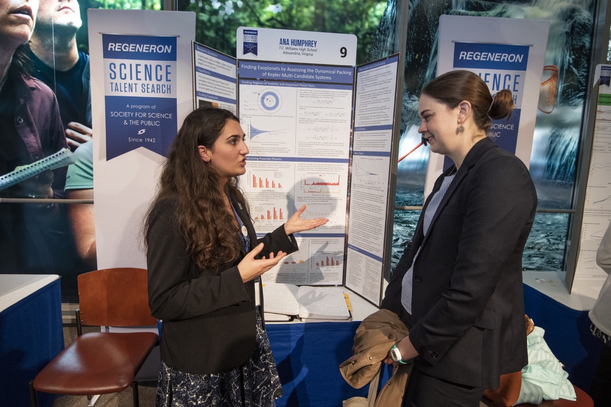 two woman speaking and gesturing with their hands in front of a poster board display of the project