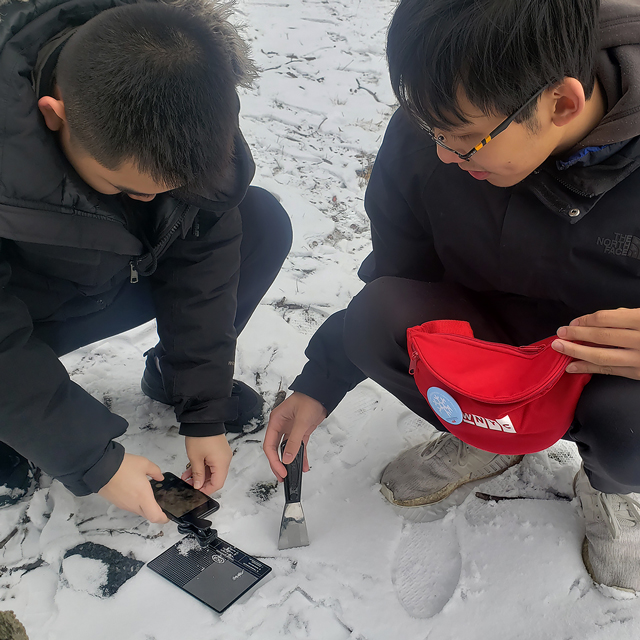 two students crouch on the snow-covered ground with the instruments from the fanny pack and inspect the snow