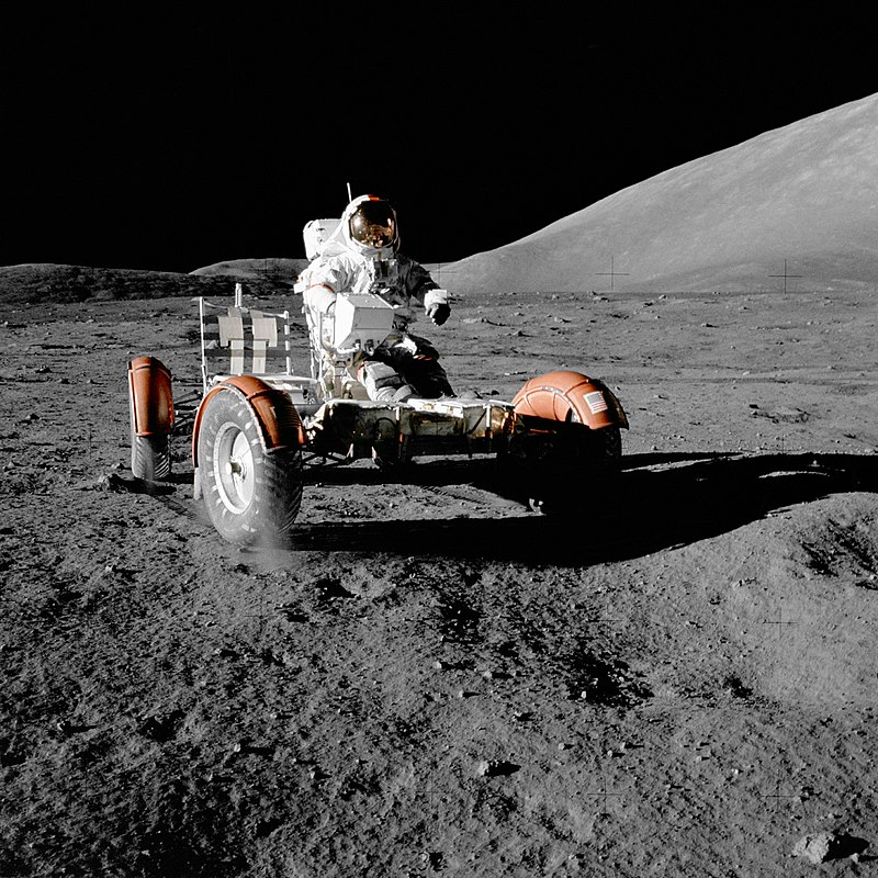 person in space suit riding a vehicle across the rocky gray surface of the moon against black sky