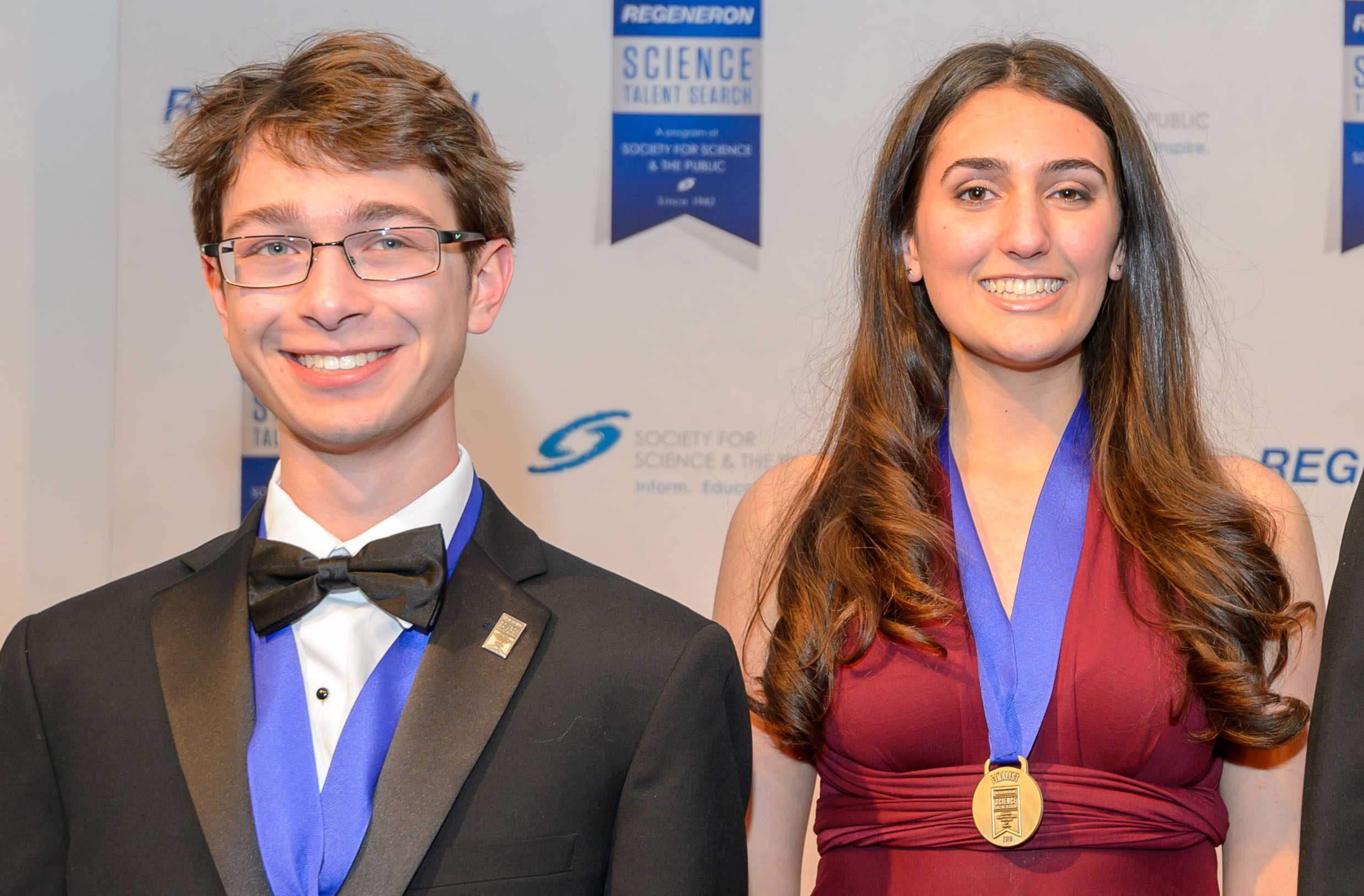 teenage boy with blond hair and wearing a bow tie standing next to teenage girl with long brown hair and red dress. both are wearing medals around their neck and smiking 