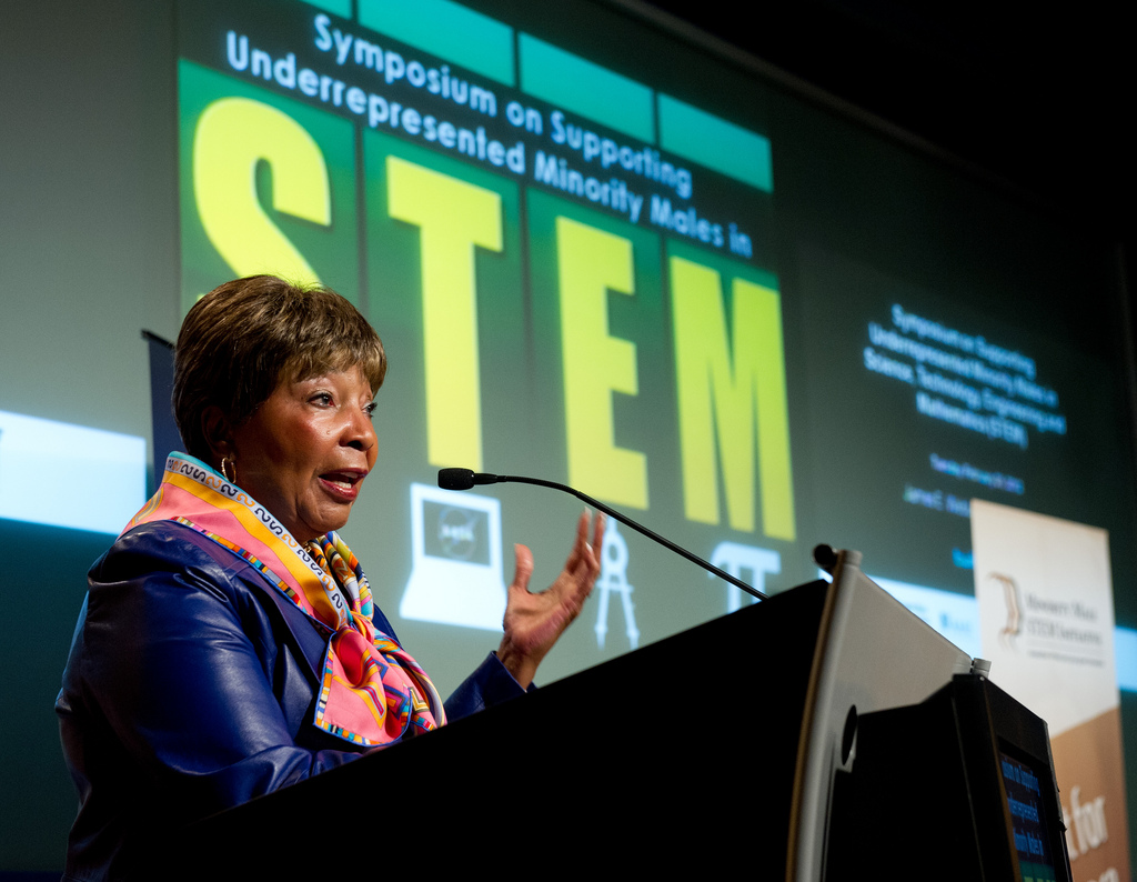 an older black woman in a brightly colored scarf and blue jacket stands at a podium giving a speech. behind her on a projection screen is "symposium on supporting underrepresented minority males in stem"