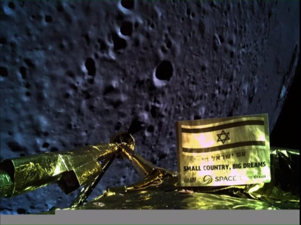 at the bottom of the image is the golden top of the beresheet lunar lander. in the background is the looming, cratered lunar surface. in the foreground of the camera, mounted on the lander, is a small golden sign that features the israeli flag and the words "Small Country Big Dreams" in both English and Hebrew