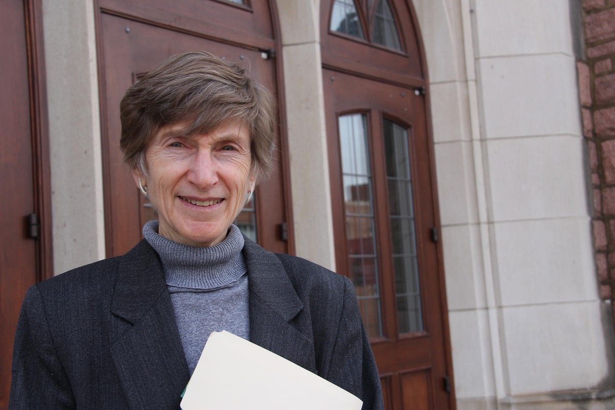 lipeles, wearing a turtleneck and blazer holding a document in front of a building