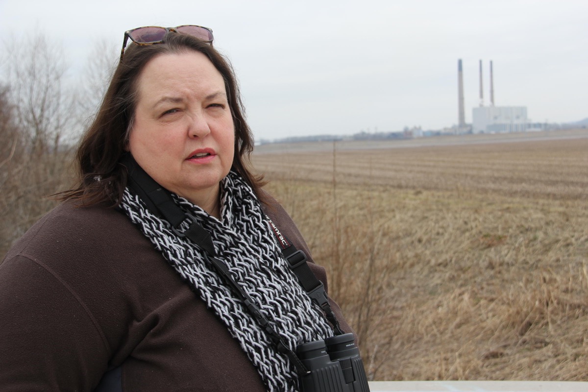 schuba stands in a field with a coal plant in the background, wearing sunglasses and binoculars around her neck