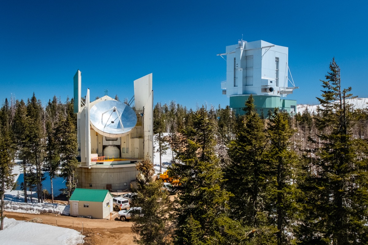 a radio dish out in the middle of a foresty mountain side covered in snow under a clear blue day