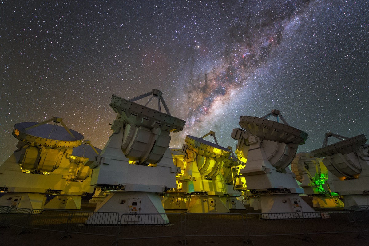 several radio dishes that are glowing softly under green-ish light. above is the night sky and the milky way galaxy