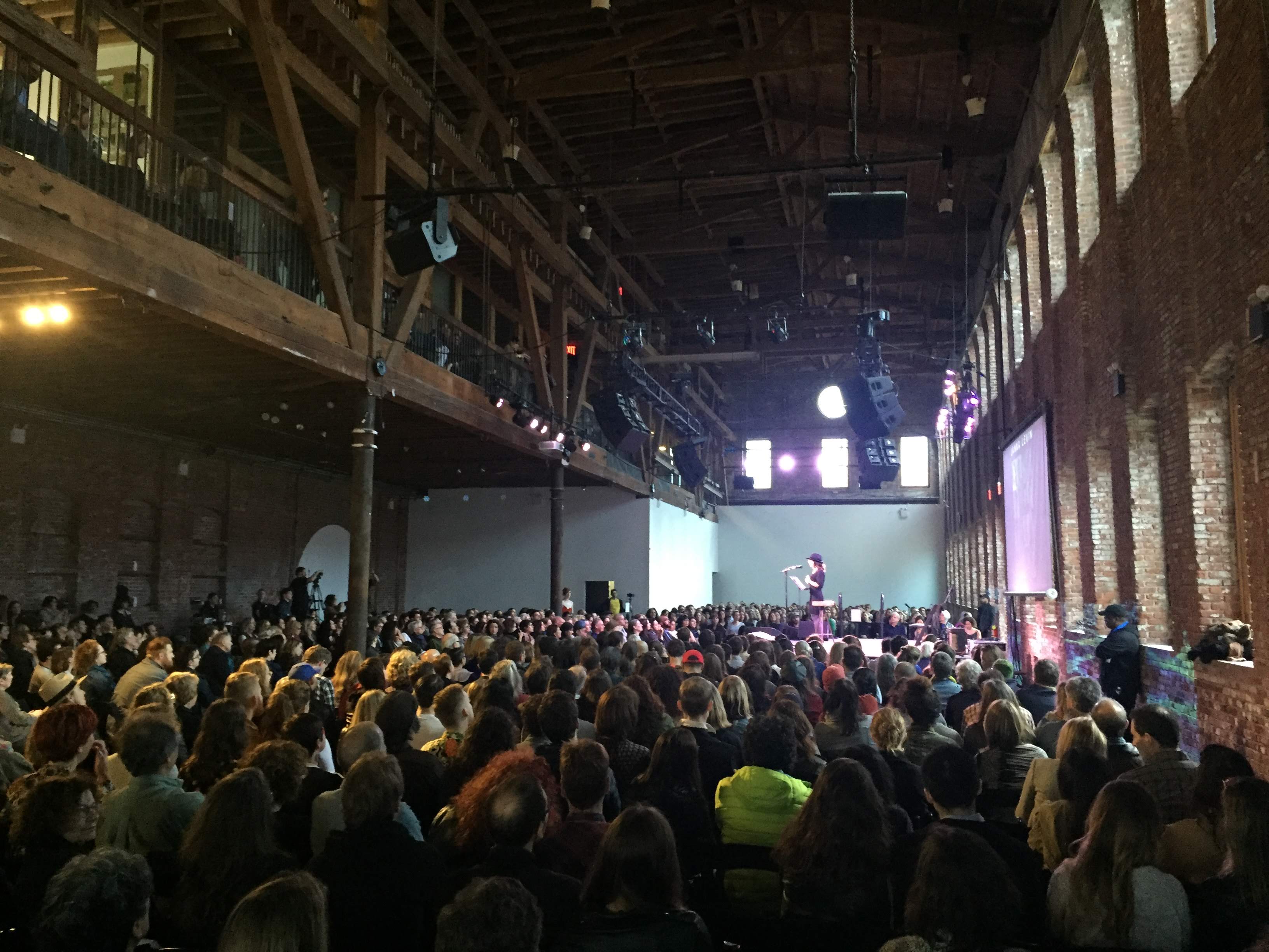 a woman on stage with a mic in a large industrial auditorium space with scores of people watching her