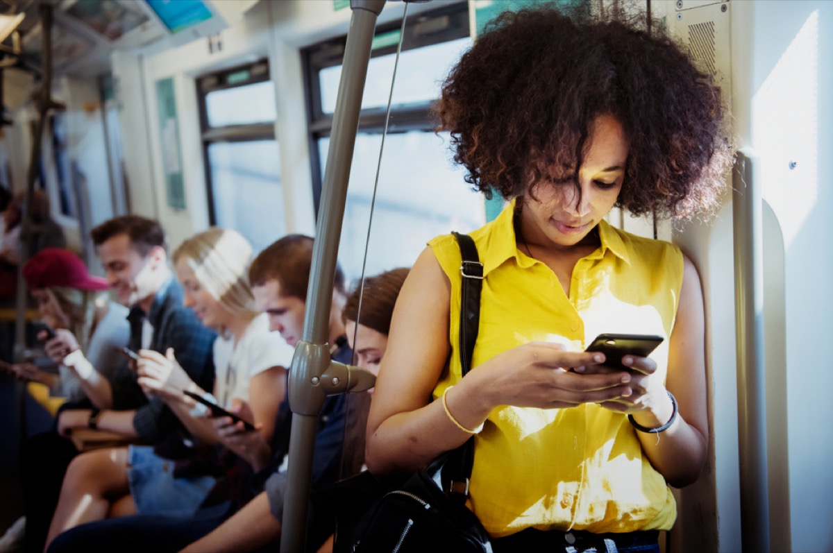 a young black woman in a yellow shirt using a smartphone on a subway train. in the background other passengers are using their phones